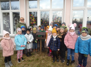Grupa dzieci z panią Arletką stoją wokół karmnika na dworze.